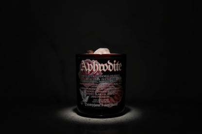 A red jar candle labeled 'Aphrodite' with rose quartz crystals on top, set on a marble surface.