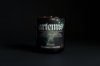 A black candle with a label featuring the name 'artemis' and an illustration of a deer. The candle is branded as 'Persephone's Sanctuary' and has various stones and plants depicted around it.