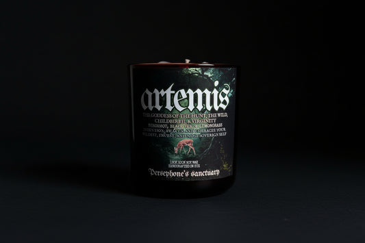 A black candle with a label featuring the name 'artemis' and an illustration of a deer. The candle is branded as 'Persephone's Sanctuary' and has various stones and plants depicted around it.