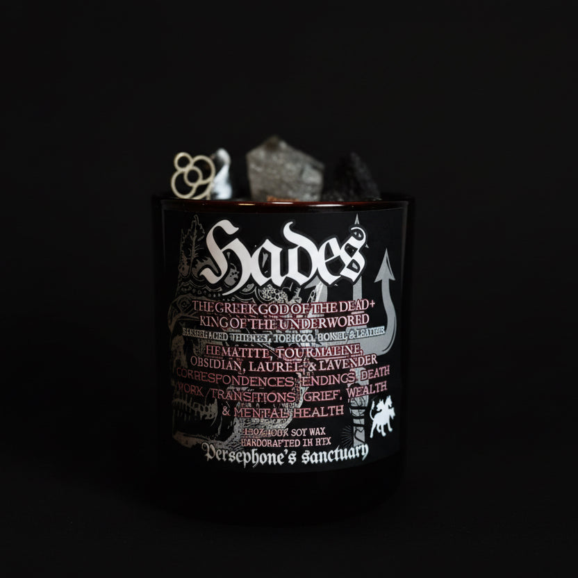 A black candle jar with 'Hades' text and mentions of components like hematite, obsidian, and bay leaf. The jar contains candles with various tops, including skull-shaped ones.