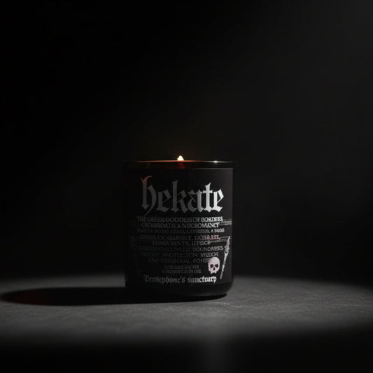 A black candle with 'hekate' written in white text, adorned with crystals and placed on a marble surface.