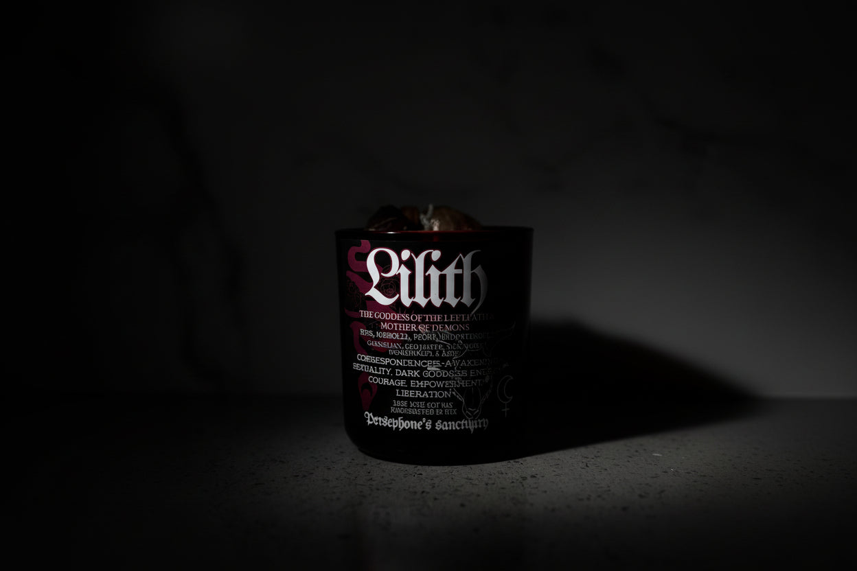 A black candle with the name 'Lilith' written on it, with a wooden wick and decorative botanical illustrations. It is set against a marble background.