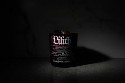 A black candle with the name 'Lilith' written on it, with a wooden wick and decorative botanical illustrations. It is set against a marble background.