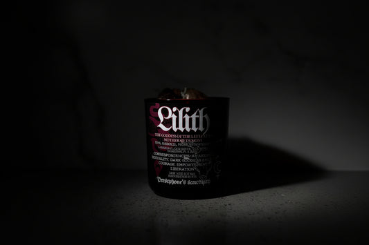 A black candle with the name 'Lilith' written on it, with a wooden wick and decorative botanical illustrations. It is set against a marble background.