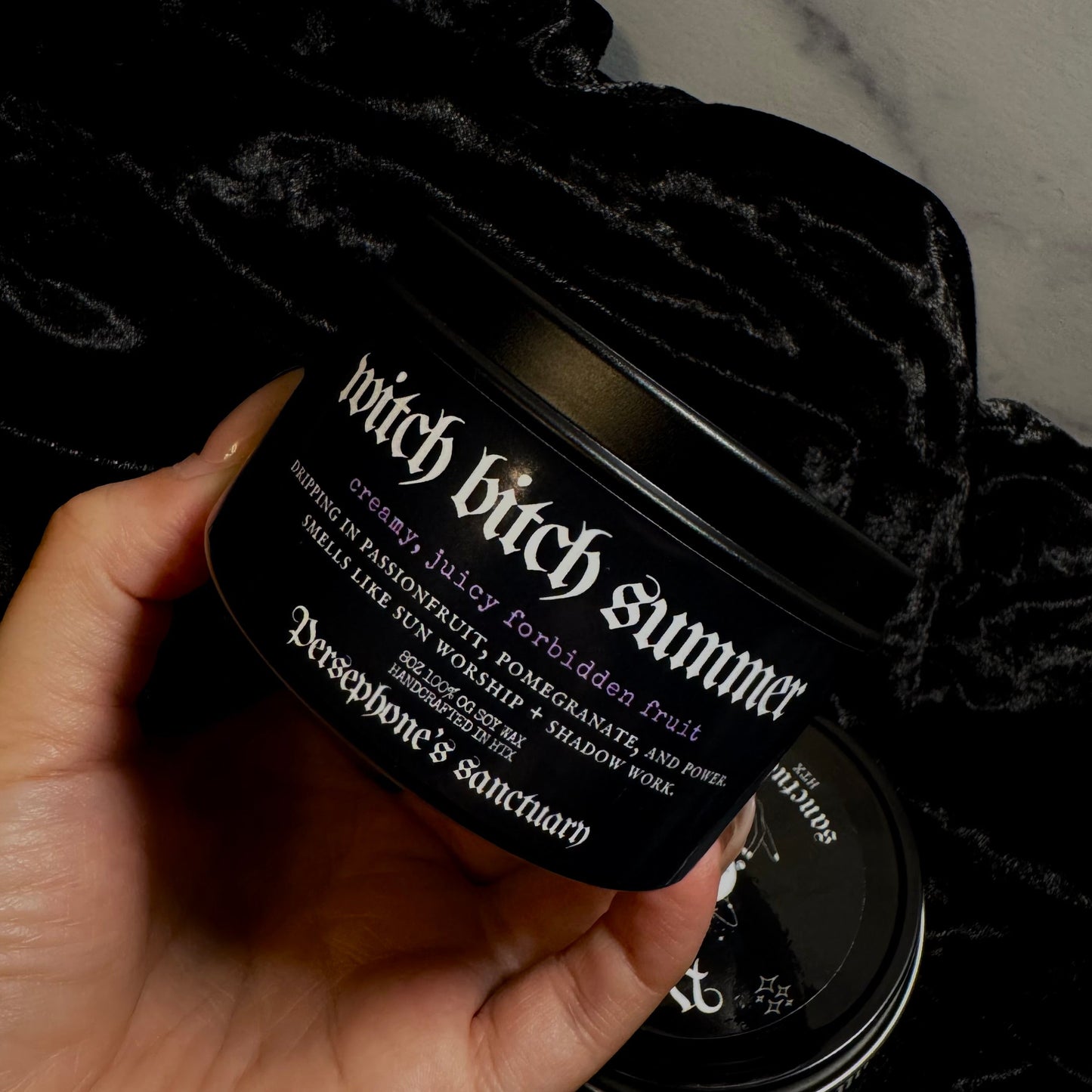 Hand holding a black candle labeled 'witch bitch summer' against a dark background.