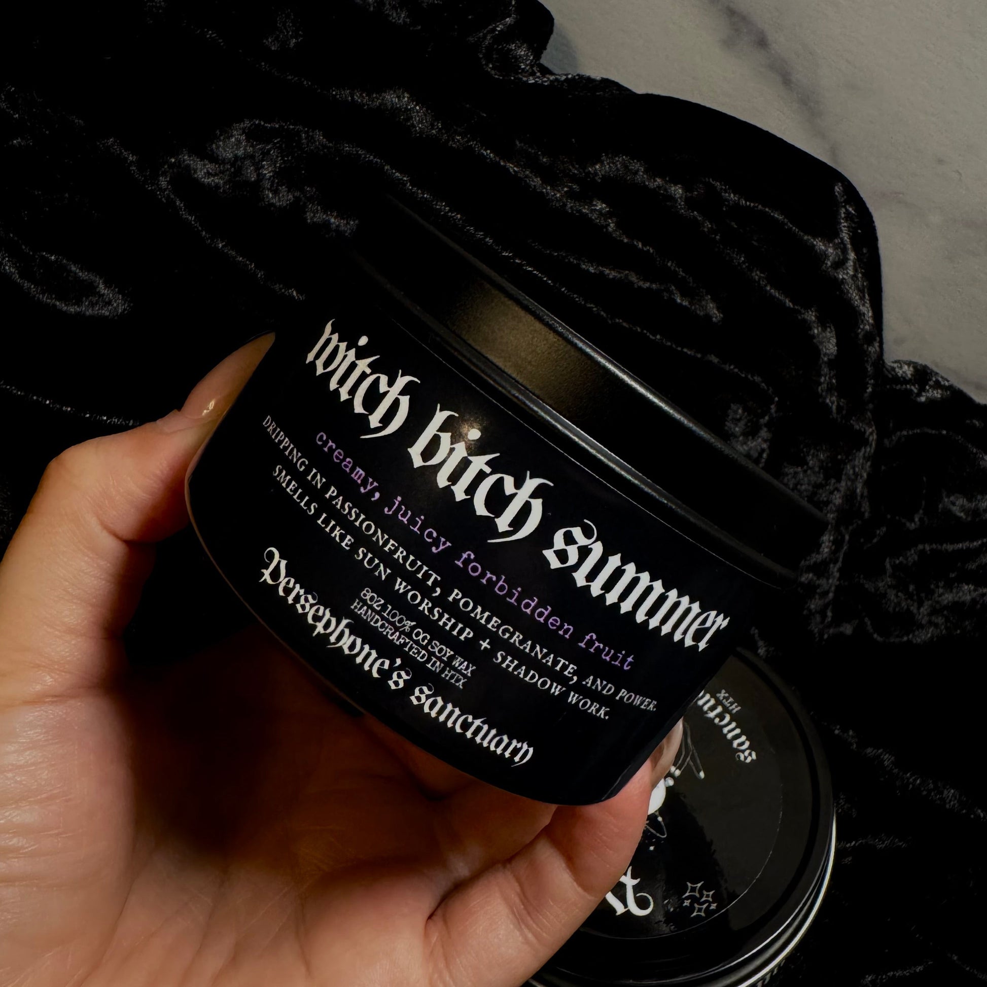 Hand holding a black candle labeled 'witch bitch summer' against a dark background.